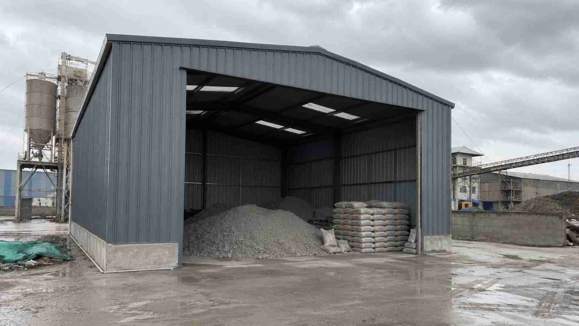 Steel-grey storage shed holding bulk cement material and stacked bags at an industrial plant.