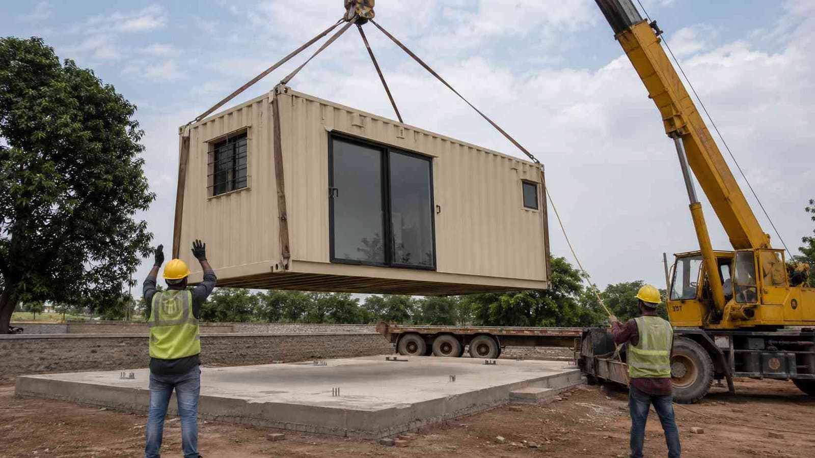 Crane lowering a Sandstone Beige two-storey inexpensive container home onto a prepared RCC plinth at a rural Indian site.