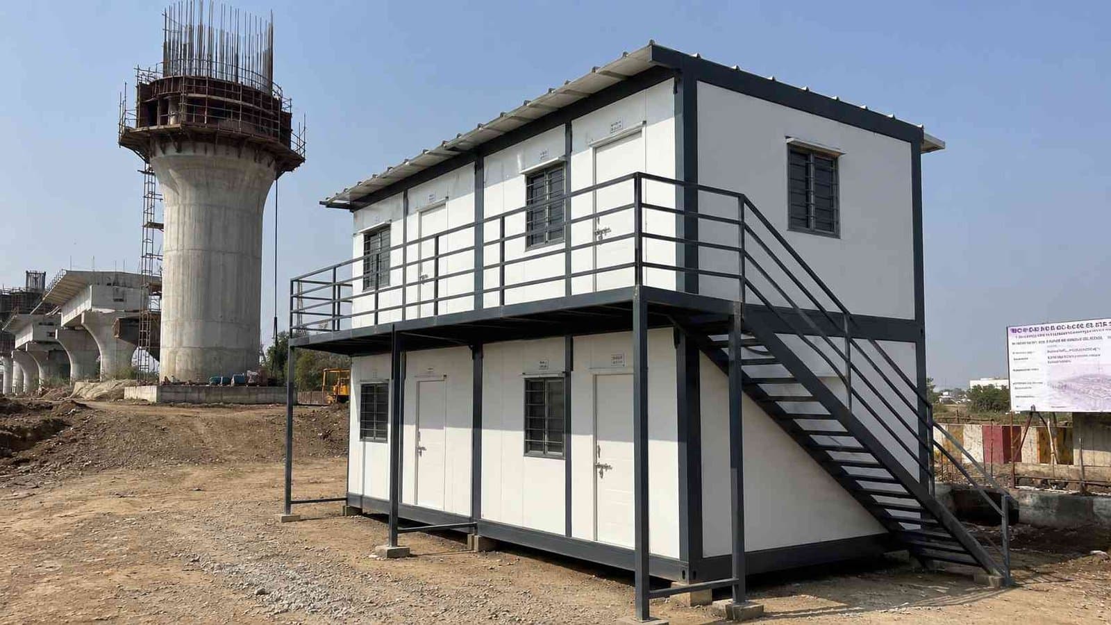 Two-tier SAMAN labor hutment block in Slate Charcoal beside a metro rail viaduct under construction.