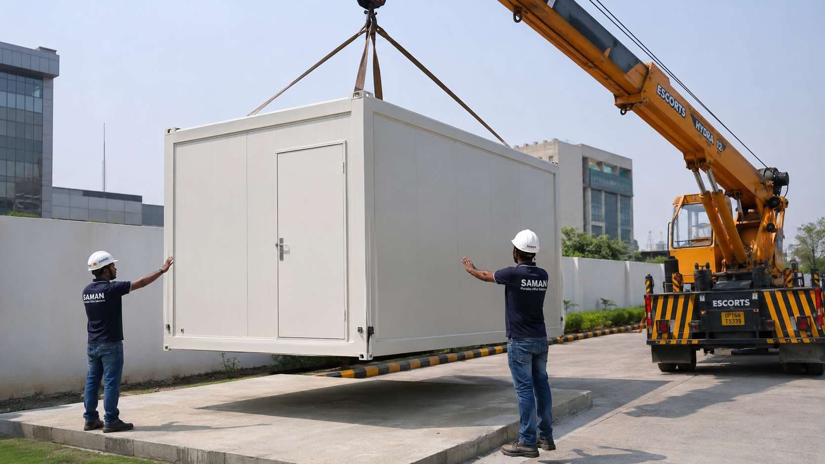 A configured modular portable office cabin being lowered by a hydra crane onto a prepared concrete pad at a Greater Noida business compound, guided by two SAMAN technicians.