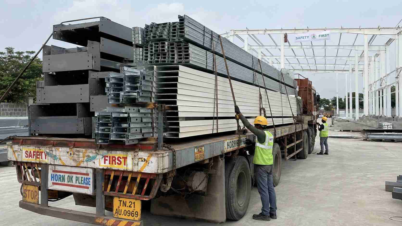 Flatbed truck loaded with pre-engineered building frame members, purlins and white cladding sheets at a dispatch yard.