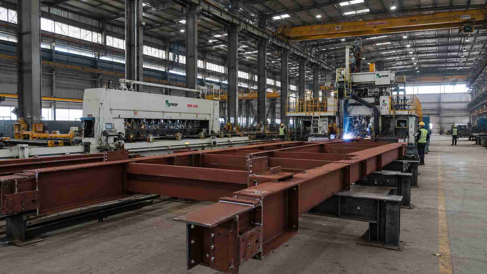 Steel fabrication plant with red-oxide primed pre-engineered building portal frames on jigs beside a CNC drilling line.