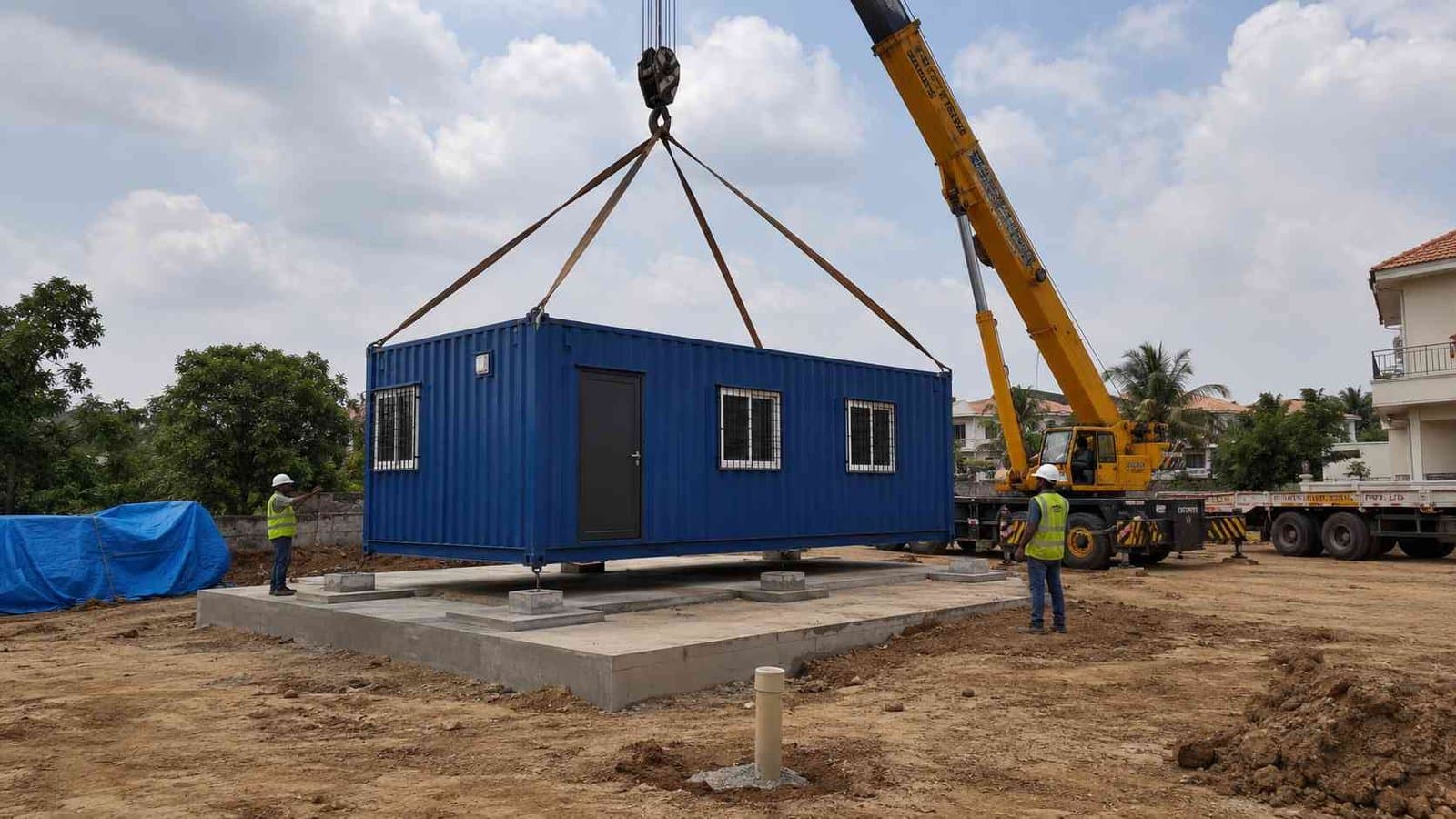 Mobile crane lowering a SAMAN Royal Cobalt prefab container home onto a prepared concrete plinth on an Indian residential site.