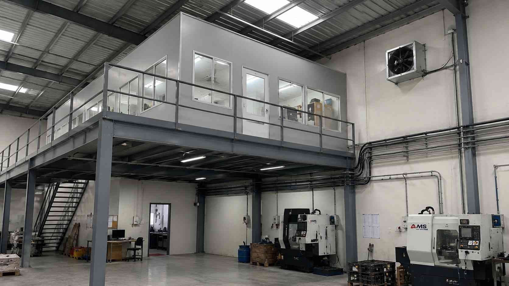 Grey steel mezzanine and wall-mounted power and ventilation inside a prefab manufacturing building.