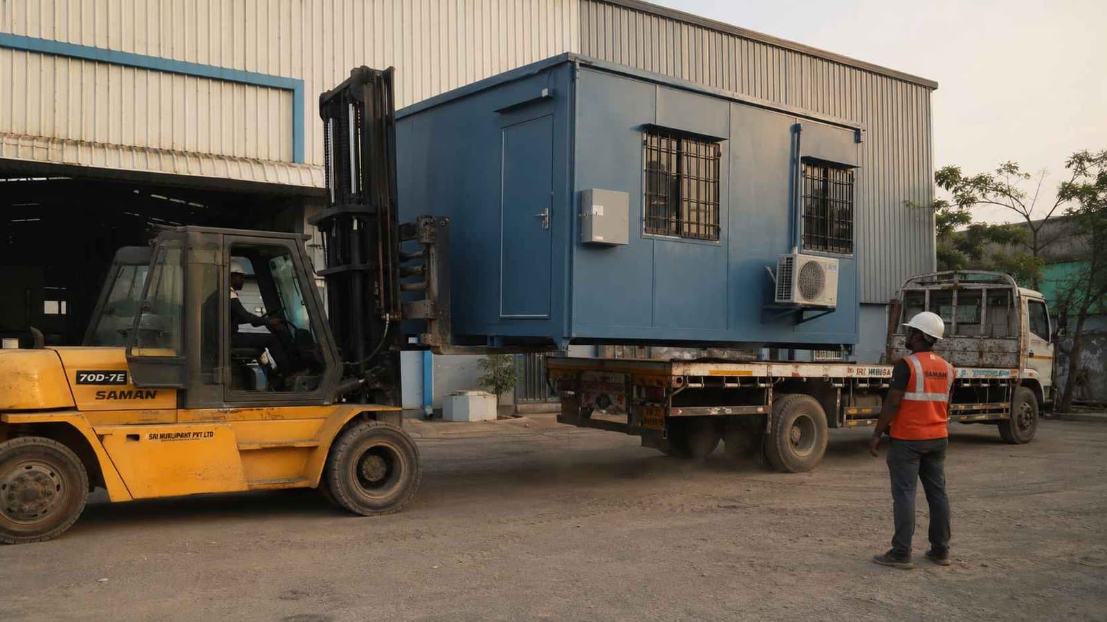 Steel Blue SAMAN cabin being loaded onto a flatbed by an industrial-yellow forklift outside an Indian warehouse.