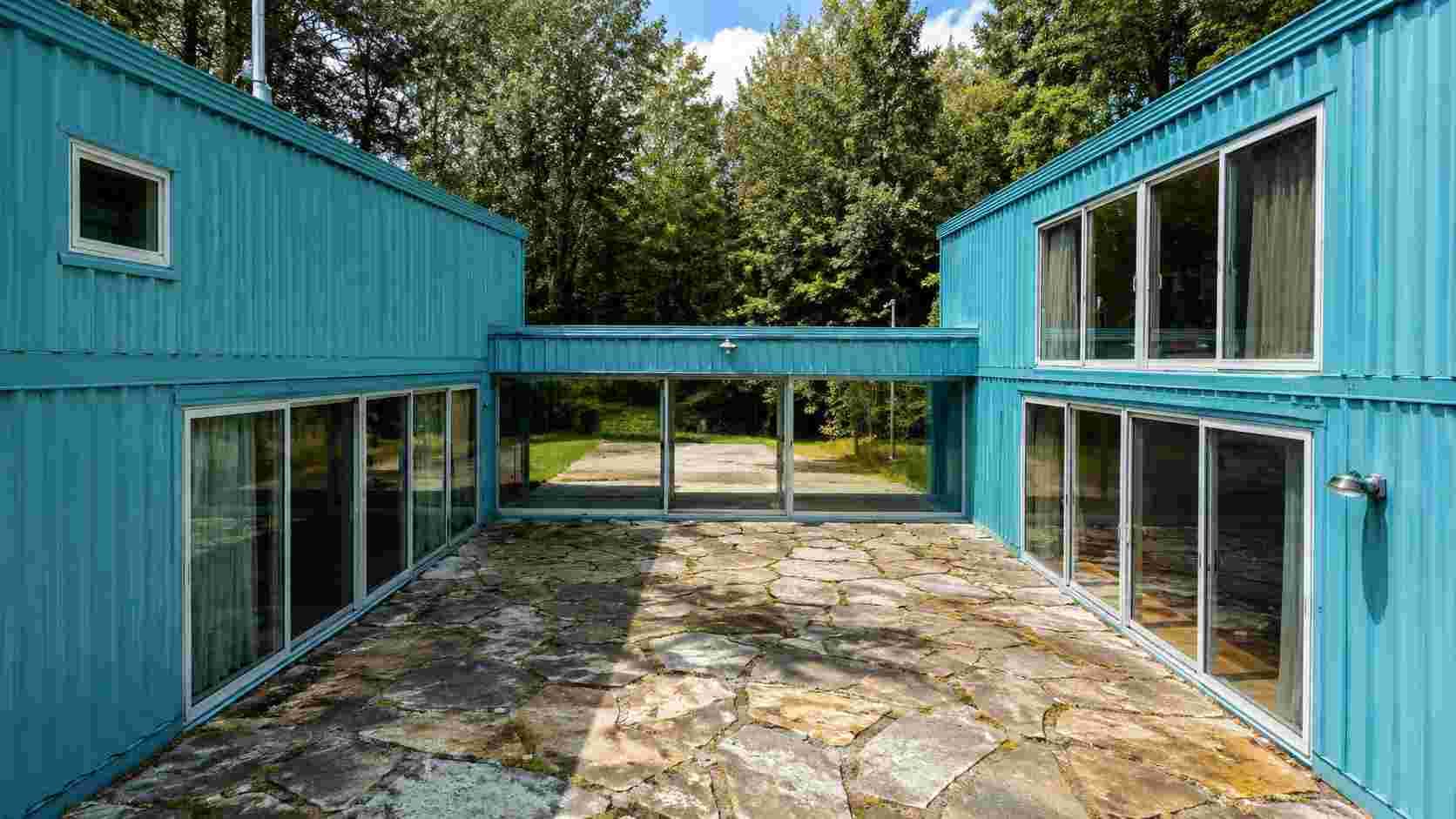 Two teal shipping container units joined around a stone courtyard with forest behind