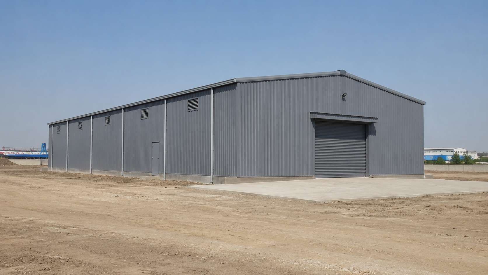 A steel-grey prefabricated warehouse standing complete on a levelled industrial plot under a clear sky.