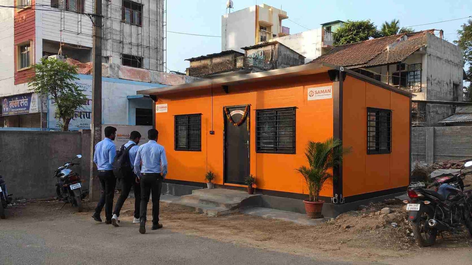 Custom orange SAMAN prefab office on a small owned plot with parked two-wheelers and arriving team.