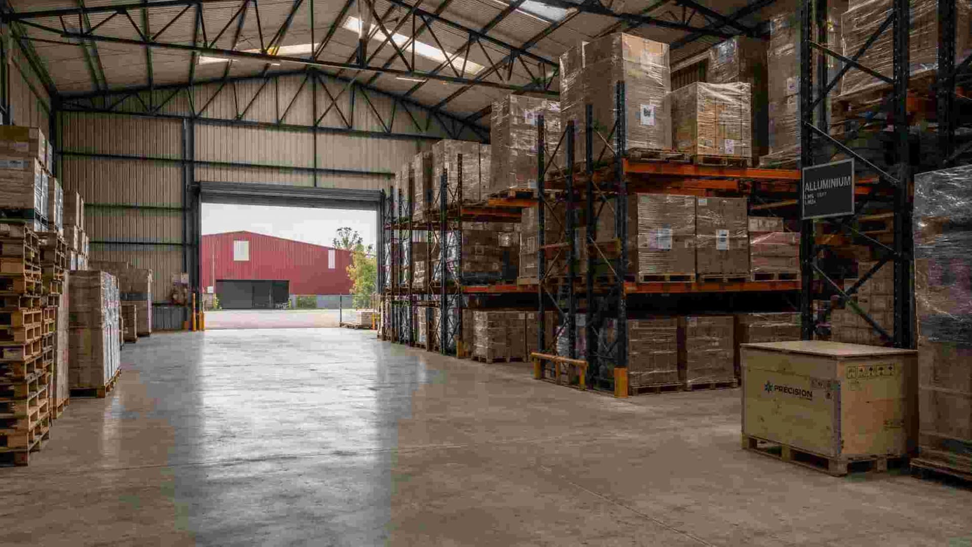 Interior of a burgundy storage shed with steel racking holding palletised goods under a steel roof.