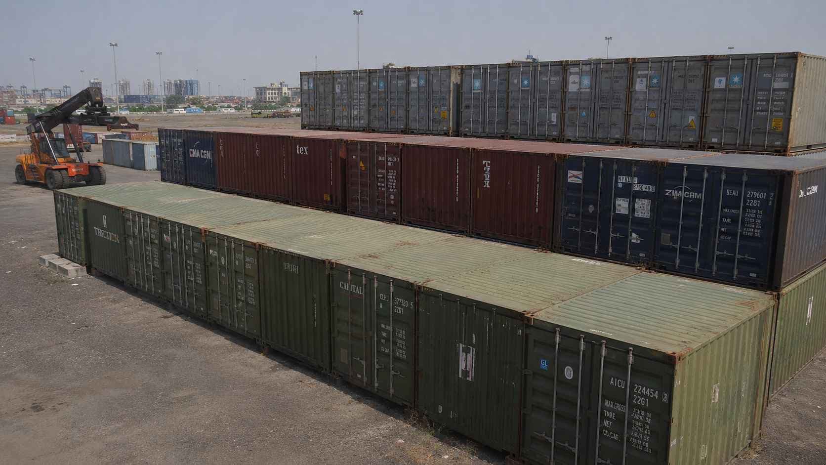 Stacked rows of used shipping containers in olive-drab and mixed colours at a major Indian port yard, with a reach stacker beside.