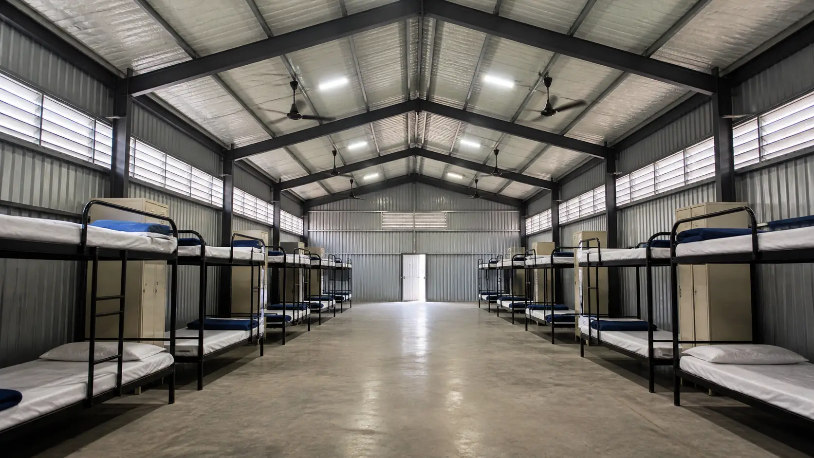 Two bunk rows facing a wide central concrete aisle inside a labour shed, with ceiling fans above